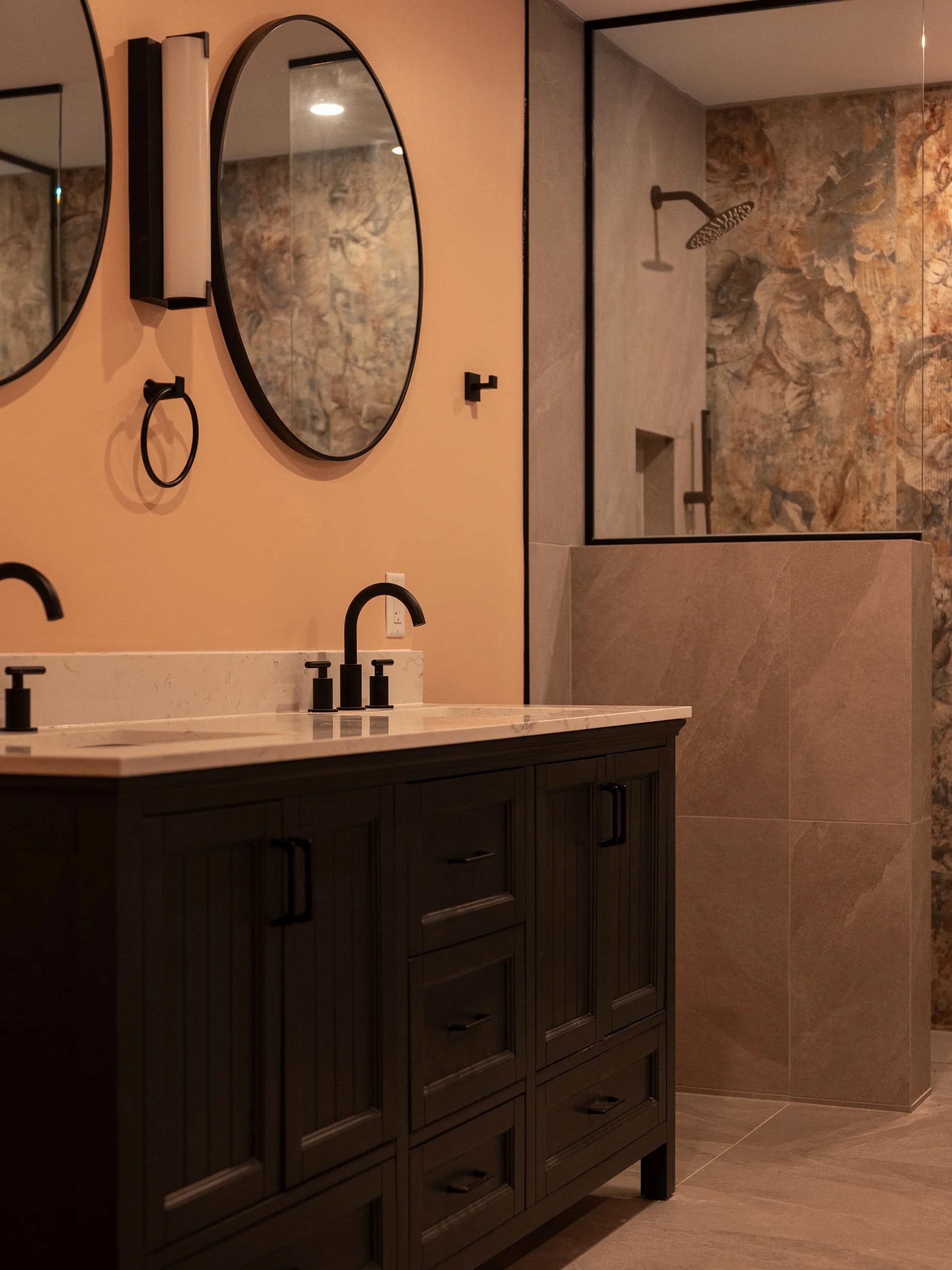Master bathroom with dark wood vanity, dual round mirrors, brass and matte black fixtures, and watercolor feature tile.